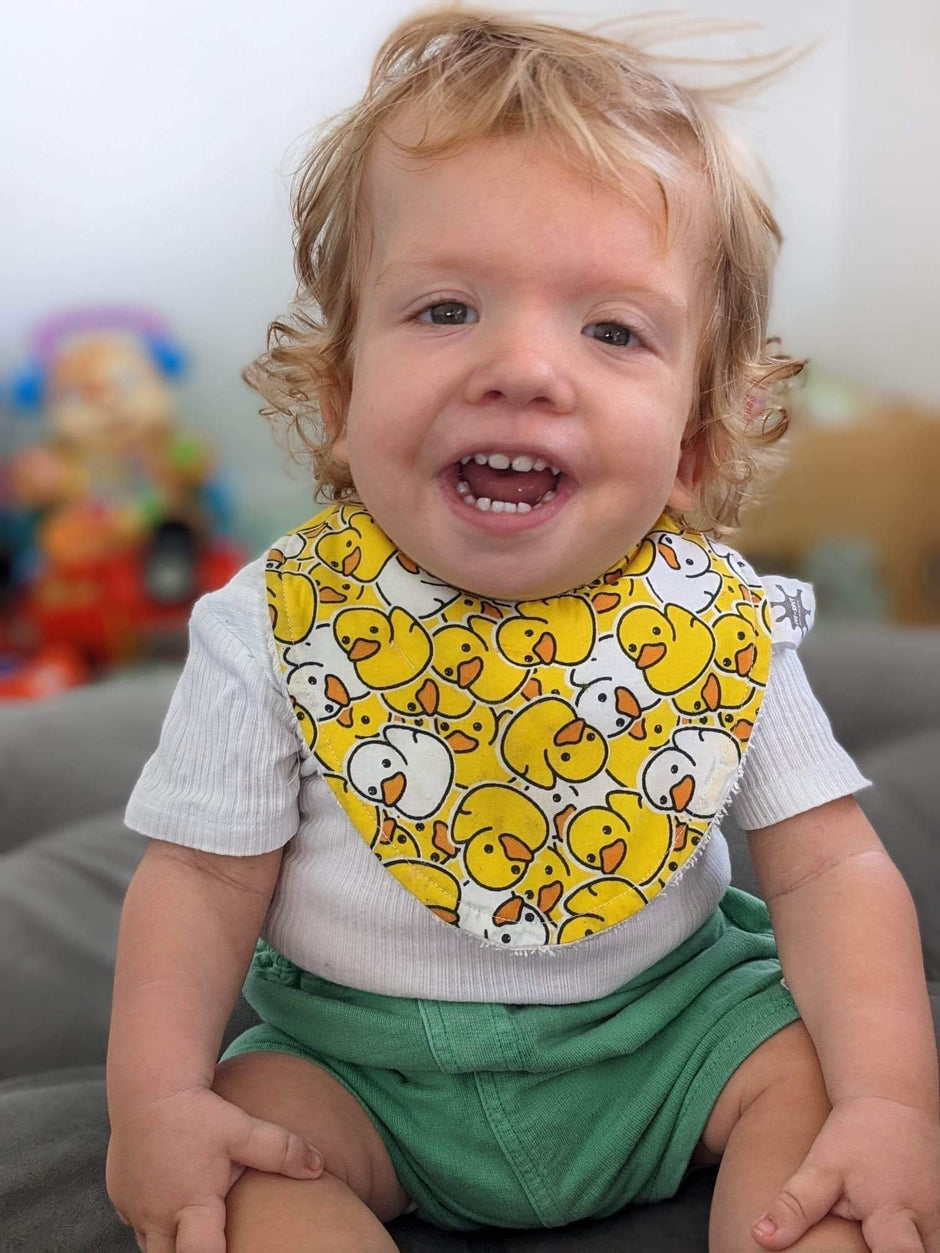 Smiling toddler wearing a bright yellow bib with a playful duck pattern, sitting happily indoors with colourful toys in the background.